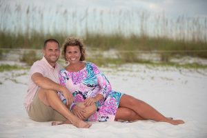 Couple smiling lounging on the beach with sea oats behind them.