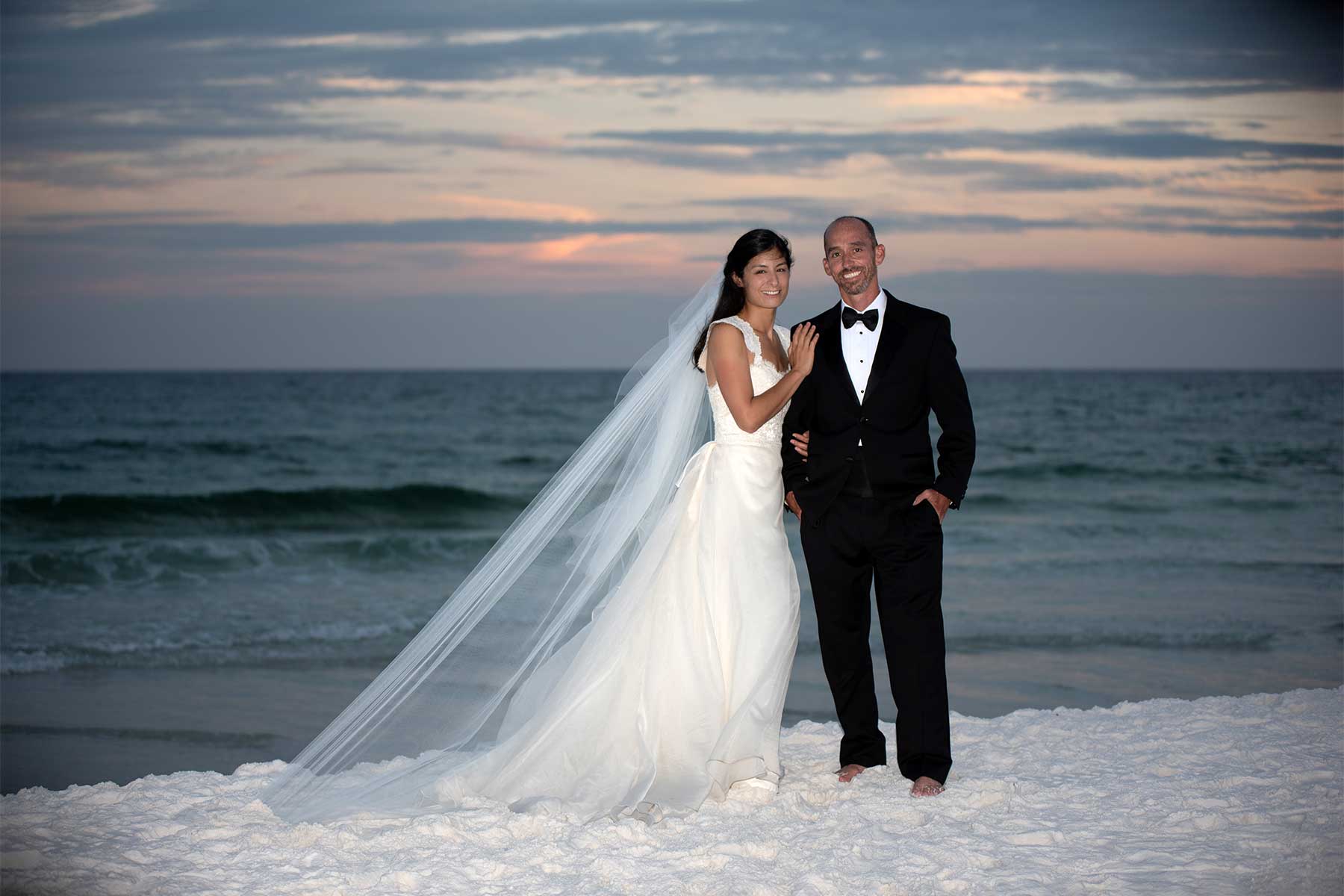A bride and groom in formal attire on the beach at sunset