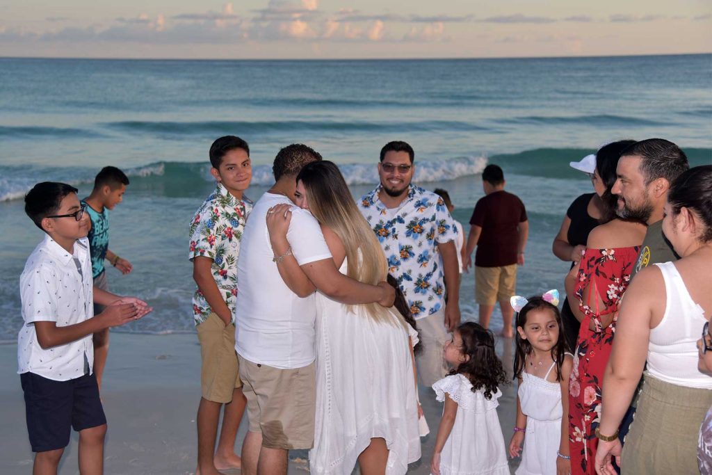 This surprise beach proposal photo shows the man and woman hugging while the family looks on.